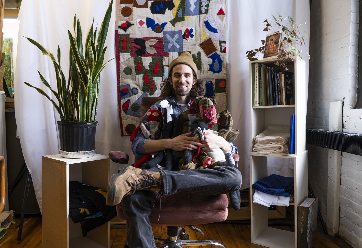 Alex Kalil in his studio holding an armful of hand-stitched soft sculptures, seated before a large patchwork quilt.